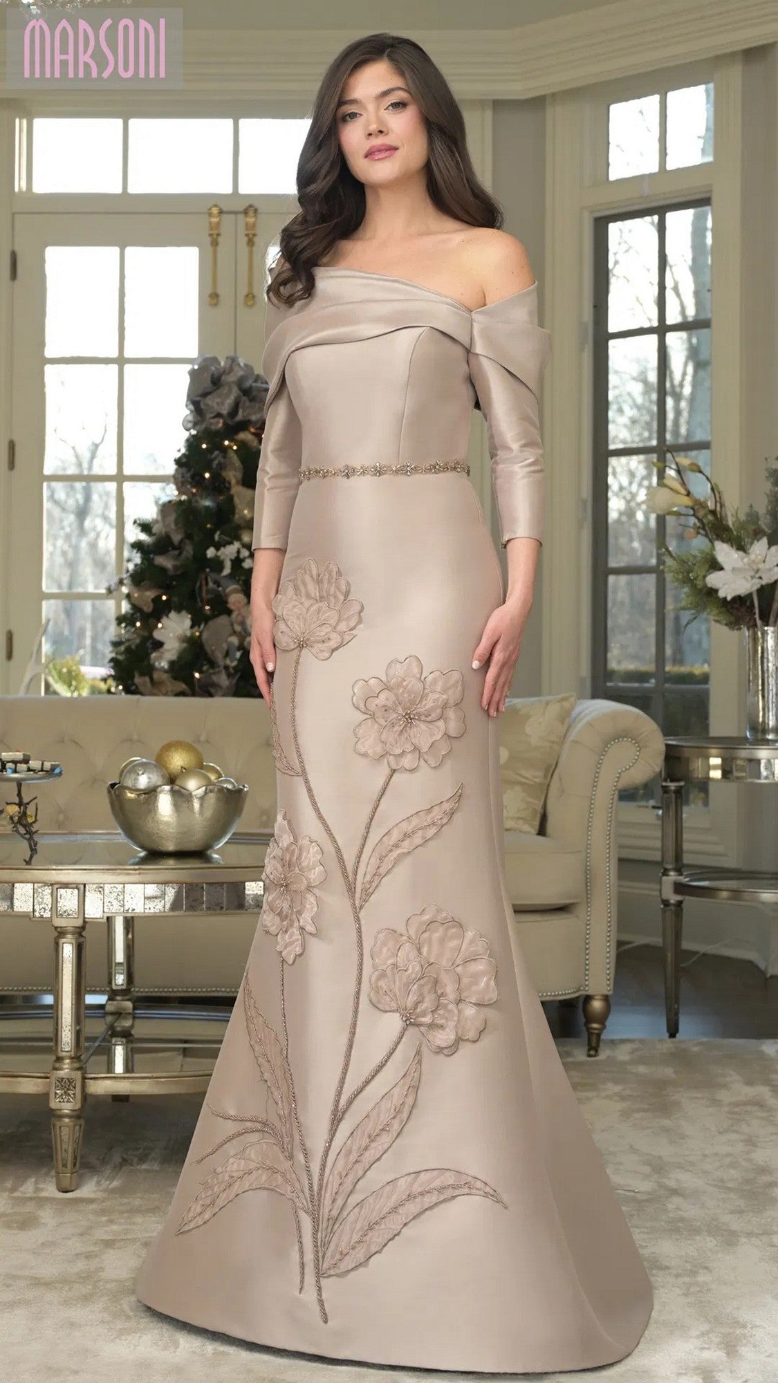 A woman stands indoors by a Christmas tree in the taupe Marsoni MV1374 long formal dress, featuring an off-shoulder neckline, three-quarter sleeves, and large floral embroidery on the fitted mermaid skirt.